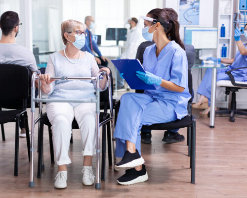 Handicapped senior woman with walking frame in hospital waiting room using walking frame talking with medical staff about treament for illness during coronavirus pandemic.